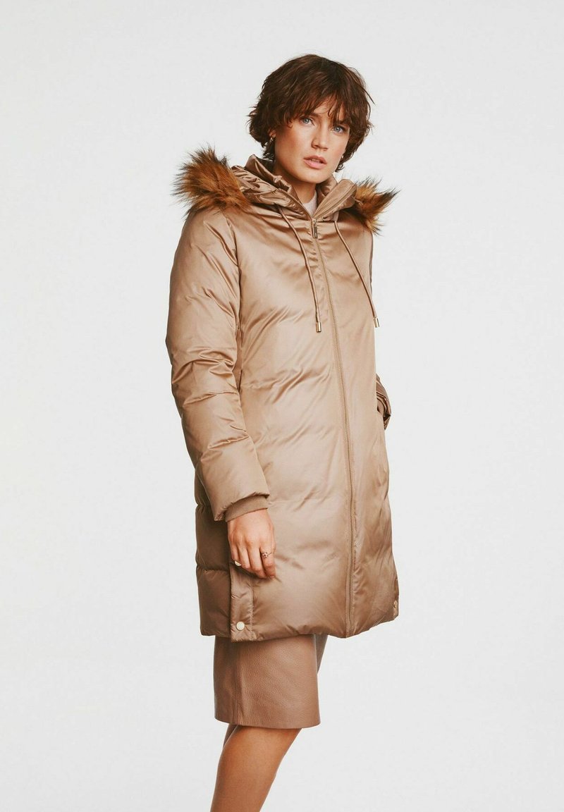 Długi beżowy płaszcz puchowy z kapturem z sztucznego futra, z zamkiem błyskawicznym i bocznymi kieszeniami. Posiada elastyczne mankiety i prosty krój.