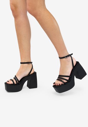 Sandalias de plataforma negras con múltiples tiras finas, un tacón cuadrado y una correa en el tobillo. Material sintético suave con un acabado mate.