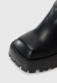 Bottine en cuir noir avec un bout carré, surface texturée, semelle en caoutchouc épaisse et motif de semelle prononcé.