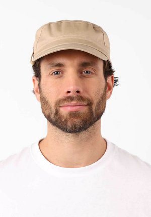 Homme avec une barbe brune et des yeux bleus, portant une casquette beige et une chemise blanche, face à un fond blanc.