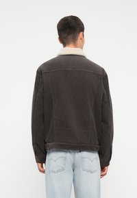 Tumehallist corduroy-tagist valmistatud jakk, millel on kreemjas kunstkarusnahast krae, nööbid ja lühike disain. Seljas on helesinised denim-püksid.