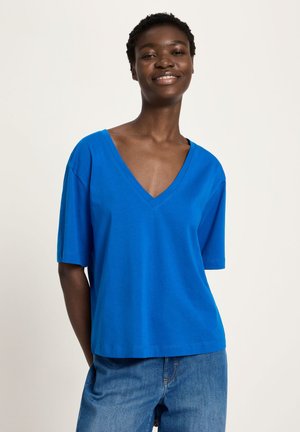 Smiling woman wearing a blue V-neck short-sleeve shirt and blue jeans, standing against a plain light background.