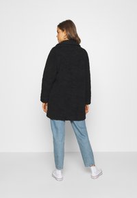 Manteau noir en fourrure avec un col, des manches longues et une coupe décontractée, associé à un jean bleu clair et des baskets blanches.