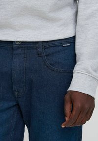 Jeans de denim en azul oscuro, con bolsillos estándar, costuras acolchadas y una etiqueta negra en la cinturilla, combinados con un sudadera gris.