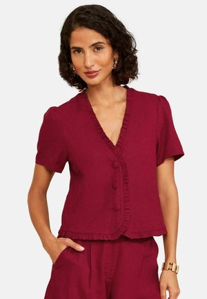 Vrouw met kort krullend haar, gekleed in een dieprode blouse met korte mouwen, knopen en ruches, gecombineerd met bijpassende broek.
