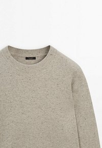 Pull beige col rond avec motif noir moucheté, col côtelé et une petite étiquette de marque noire visible à l’intérieur du col.