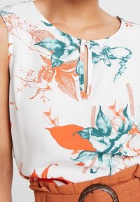 Bezrukává blůza z bílé látky s živým oranžovým a tyrkysovým květinovým potiskem. Obsahuje výstřih ve tvaru klíčové dírky a hladkou texturu.