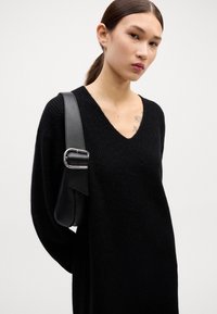 Pulover negru supradimensionat cu decolteu în V, dintr-un material tricotat texturat. O geantă neagră cu un mâner lat și detalii din inox se află pe umăr.