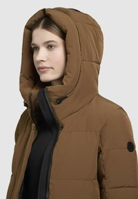 Barna puffdzseki nagy kapucnival, fekete cipzárral és nyomógombokkal, sima textúrával és logóhoz hasonló folttal a számnál.