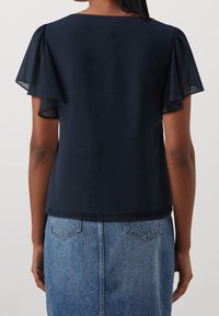 Blouse bleu marine avec manches volumineuses transparentes, tissu lisse et encolure arrondie, associée à une jupe en jean.