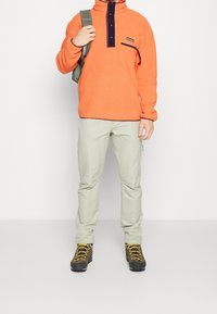 Hombre con un pulóver de forro polar naranja brillante, pantalones beige claros, botas de senderismo marrones con cordones amarillos, sosteniendo la correa de una mochila verde sobre el hombro.
