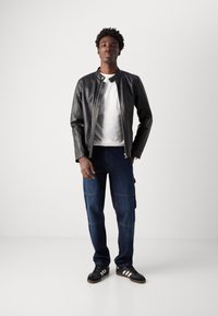 Black leather jacket, white t-shirt, blue cargo pants with pockets, and black sneakers with white stripes. Model stands in a neutral background.