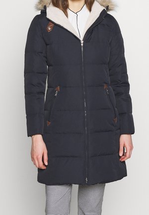 Femme portant un long manteau d'hiver noir matelassé avec capuche bordée de fourrure, poches zippées et fermeture éclair à l'avant, sur un pantalon à carreaux noir et blanc.