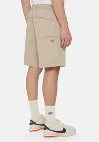 Beige shortsit, joissa on joustava vyötärö, kaksi takataskua ja pieni logo sivussa. Yhdistettynä valkoisiin sukkiin ja harmaisiin lenkkareihin.