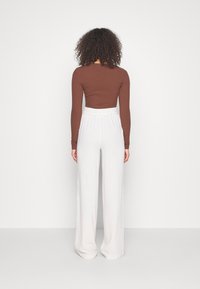 Kvinna med lockigt hår står vänd mot en vit vägg, iklädd en brun långärmad tröja och vita högmidjade vida byxor.