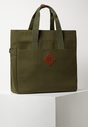 Sac fourre-tout en toile vert olive avec doubles poignées supérieures, une poche avant et un patch en cuir marron en forme de losange à l'avant.