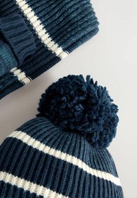 Blå strikket beanie med en stor pom-pom på toppen, med kontrasterende hvide striber i et struktureret mønster. En anden lignende hat er delvist synlig.