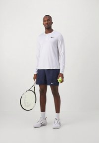 Weißes langärmeliges Sportshirt, blaue Shorts, hält einen Tennisschläger und Ball. Nike-Logo auf Shirt und Shorts. Weiße Turnschuhe.
