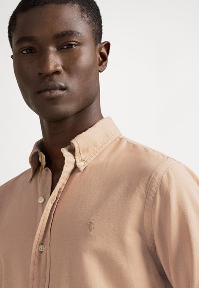 Jauns vīrietis valkā gaiši brūnu kreklu ar pogām un apkakli ar pogām, stāvot pret vienkāršu baltu fonu.