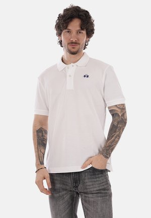 Uomo con capelli ricci e tatuaggi su entrambi gli avambracci, indossa una polo bianca e jeans grigi, in piedi con una mano in tasca.