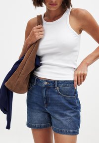 Beli rebrasti top brez rokavov v kombinaciji z modrimi denimsko kratkimi hlačami. Kratke hlače imajo klasičen kroj z vidnimi šivi in gumbnim pasom.