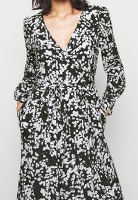 Mustavalkoinen mekko, jossa on valkoinen kukkakuvio, V-kaula-aukko, pitkät hihat, vyötärönauha ja sivutaskut, valmistettu kevyestä kankaasta.