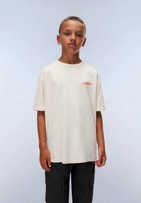 Valkoinen puuvillapaita, jossa on pyöreä kaula-aukko ja lyhyet hihat, ja rinnan kohdalla on hillitty oranssi graafinen kuvio. Yhdistetty mustiin cargo-housuihin.