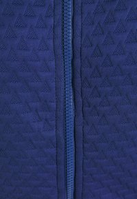 Marinblå zip-up plagg med ett texturerat triangulmönster, med en blå dragkedja i mitten. Slät tyg med en strukturerad utseende.