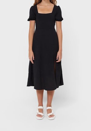 Zwarte midi-jurk met een vierkante halslijn, korte pofmouwen, zijsplitten en een gladde textuur. Gecombineerd met witte sandalen.
