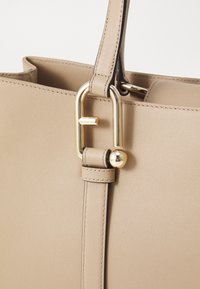 Sac à main en cuir beige, de forme rectangulaire, avec une anse supérieure et des accessoires en métal doré, ornés d'un logo distinctif.
