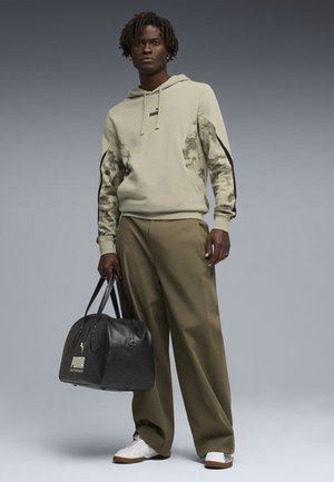 Uomo che indossa una felpa con cappuccio beige con maniche mimetiche, pantaloni verde oliva a gamba larga, sneakers bianche, con una borsa da palestra nera Puma, in piedi davanti a uno sfondo grigio.