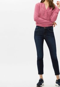 Rosa Bluse mit langen Ärmeln, V-Ausschnitt und Knopfdetails, kombiniert mit dunkelblauen Skinny Jeans und navyfarbenen Sportschuhen.