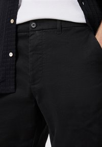 I chinos in cotone neri presentano una superficie liscia, un unico bottone nero e tasche laterali discrete, abbinati a una camicia scura con texture.