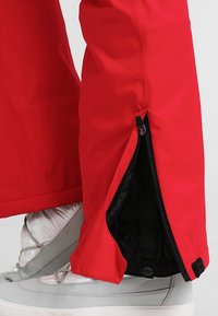 Pantalon imperméable rouge avec ouvertures à glissière aux chevilles, doté d'une texture lisse et brillante et d'une doublure noire contrastante. Des chaussures argentées sont visibles.