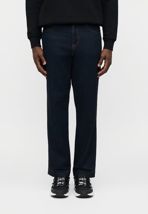 Uomo che indossa jeans dritti blu scuro, felpa nera e scarpe da ginnastica nere con lacci bianchi su uno sfondo neutro.