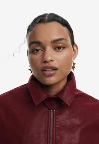 Veste en cuir bordeaux avec un col haut et une fermeture éclair argentée. Le modèle porte des boucles d'oreilles en forme d'étoile. Sa peau a des taches de rousseur et elle affiche une expression neutre.