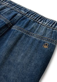 Tela de mezclilla con cinturilla elástica y costuras visibles, con un pequeño logo marrón bordado cerca del área del bolsillo.