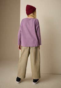 Long-sleeve lavender top, baggy olive green pants, black sneakers. Model wears a maroon knit beanie, facing away against a neutral background.