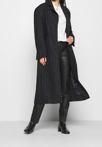 Kobieta w długim czarnym płaszczu w prążki, białej bluzce, czarnych skórzanych spodniach i czarnych botkach na obcasie, stojąca na jednolitym tle.