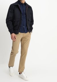 Giacca leggera blu marina con due tasche frontali e fodera a quadri, abbinata a una camicia blu marina a pois, pantaloni color cammello e sneaker bianche.