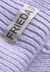 Lila gebreide stof met ribbelstructuur en een wit label met "FRIEDA" in zwarte letters, dat de zachte, donzige garen details laat zien.