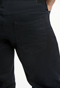 Zwarte jeans met een close-up van de achterzak, met subtiele stiksels en een gladde, zachte textuur. Minimale details en een strak ontwerp.