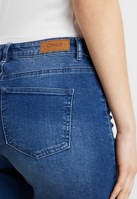 Modré džíny z denimu s koženou nášivkou s nápisem "ONLY" na zadním pásku. Tkanina má hladkou strukturu s minimálním vyblednutím.