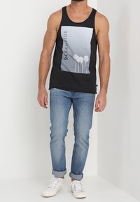Top nero senza maniche con una grafica grigia che raffigura palme, abbinato a jeans azzurri chiari e sneakers bianche. Il tessuto sembra liscio e leggero.