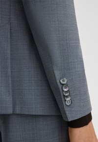 Grijze colbertmouw met een subtiele geweven textuur, voorzien van vier grijze knopen en een contrasterende donkere gebreide manchet.