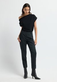 Femme portant un top noir à une épaule, un pantalon noir ajusté et des bottines à talons noirs sur un fond clair.