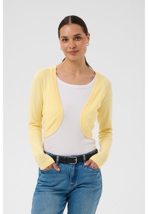 Jeune femme aux cheveux foncés attachés en queue de cheval, portant un cardigan court jaune par-dessus un haut blanc et un jean bleu, les mains dans les poches.