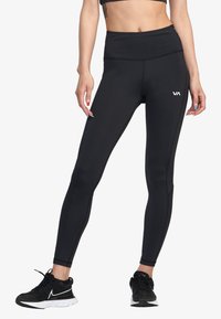 Leggings neri a vita alta realizzati in tessuto elastico con cuciture piatte e un logo sulla coscia sinistra, abbinati a scarpe da corsa nere.