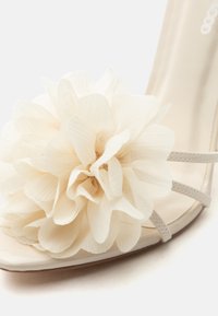 Μπεζ σανδάλι ανοιχτής μύτης με πολλαπλά λεπτά λουράκια και μεγάλο υφασμάτινο λουλούδι με υφή στο μπροστινό λουρί.