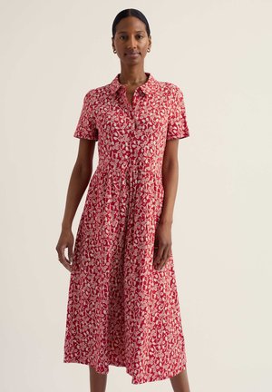 Femeie îmbrăcată într-o rochie midi roșie cu model floral alb, mâneci scurte, guler și nasturi în față, stând într-un decor simplu.